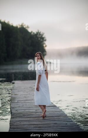 Vista posteriore di giovane donna in abito bianco in piedi da sola sul ponte pedonale e fissando al lago. Mattina fredda con una nebbia sull'acqua Foto Stock