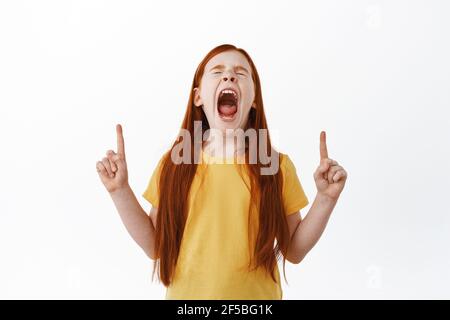 Bambina dai capelli rossi che urla e punta le dita verso l'alto. Zenzero capretto con grida e mostrando qualcosa da parte sullo spazio di copia, bianco Foto Stock