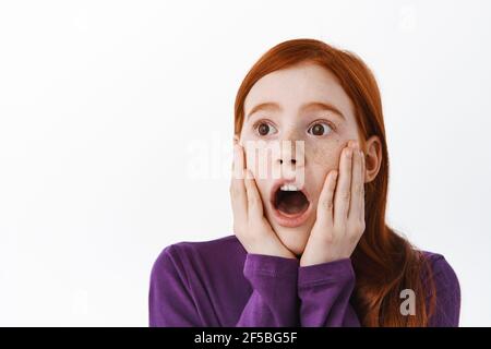 Sorprende il ragazzino rosso, ragazza con i capelli di zenzero e i fracles che si stanno facendo a pezzi, fissando da parte con il viso scioccato, in piedi su sfondo bianco Foto Stock