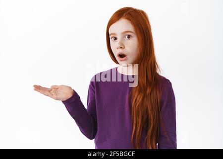 Sorpresa e stupito bambino carino, zenzero ragazza con freckles che tiene l'articolo sul palmo, mostra il tuo logo sulla mano contro copyspace, sfondo bianco Foto Stock