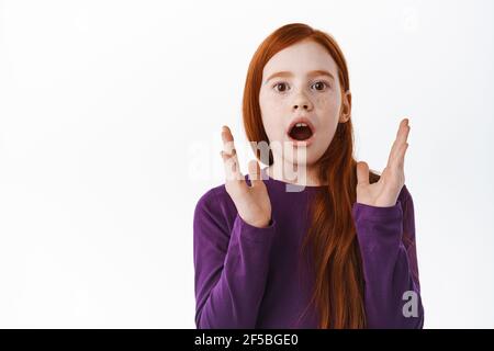 Impressionato zenzero ragazza con i greckles che sbattono, le mani clap e sguardo stupito e stupito con impressionante cosa impressionante, il controllo fuori, levandosi in piedi sopra bianco Foto Stock