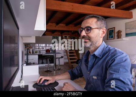 Ritratto di uomo spagnolo caucasico che lavora su computer felice, seduto in ufficio a casa. uomo di 42 anni con bicchieri e barba Foto Stock