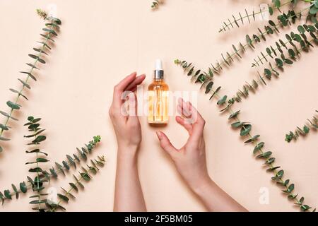 La mano femminile contiene bottiglie di vetro di essenza o siero con foglie di eucalipto naturale su sfondo beige. Cosmetici naturali e spa a disposizione Foto Stock