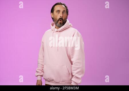 Impressionato speechless scioccato adulto bearded uomo capelli grigi in fresco pink hoodie pieghevole labbra wow occhi schioccanti macchina fotografica sorpreso stupito incuriosito Foto Stock