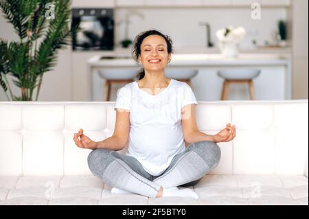 Calma bella donna di razza mista gravidanza felice, riposante mentre si siede a casa in soggiorno sul divano in posizione lotus, occhi chiusi, sorridente. La madre in attesa sta raffreddando, allevia lo stress Foto Stock