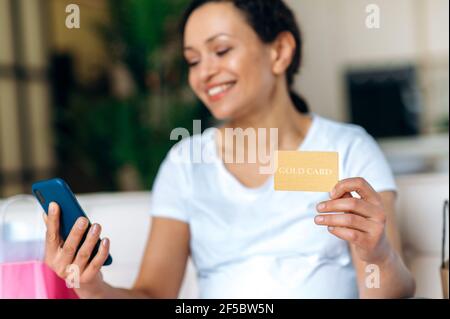 Donna di razza mista felice che indossa una t-shirt bianca con carta di credito e smartphone, shopping online, inserire un numero di carta, sorridendo. Donna effettua gli acquisti nel negozio online, o ordina la consegna a casa Foto Stock