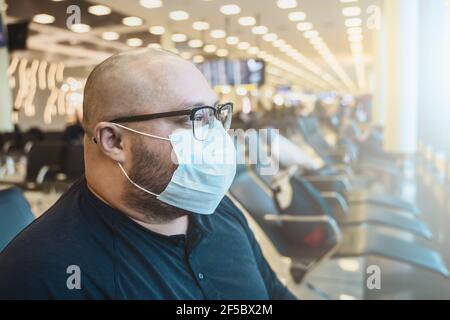 Uomo in occhiali in maschera protettiva in attesa di salire a bordo presso la sedia aeroporto terminal. Trasporto in aereo di lavoro o viaggio in covid-19 periodi pandemici. Foto Stock