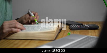 foto panoramica di una persona mano che sottolinea un libro, con un evidenziatore verde, con computer portatile, calcolatrice e cellulare, luce drammatica. giorno del lavoratore, Foto Stock