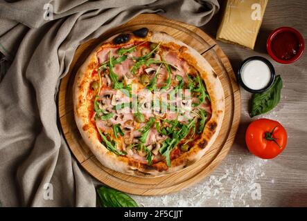 Pizza fatta in casa con rucola, sottaceti, prosciutto e salsa su un piatto di legno per pizza. Pizza sul tavolo decorata con un pezzo di stoffa, farina, formaggio, pomodoro Foto Stock