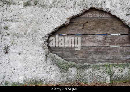 Calcestruzzo e vecchie tavole di legno. Un foro in una recinzione grigia con bordi irregolari è stato patchato con le tavole. Attraverso le fessure tra le barre. Muschio su un cemento Foto Stock