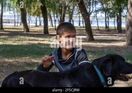 Ritratto emozionale di un ragazzo che strinse un cane nero con un collare blu nel parco. Un bambino di 10 anni e una donna di 11 anni Labrador. Una passeggiata nel Foto Stock