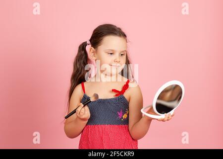 ritratto di una ragazza con i ponytail che tiene una spazzola di polvere nelle sue mani e che guarda in uno specchio bianco di plastica, isolato su uno sfondo rosa Foto Stock