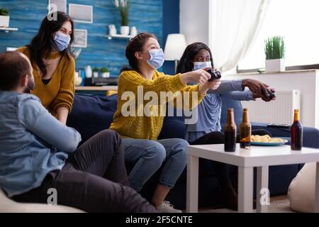 Amici di corsa misti che si incontrano per giocare a videogiochi con controller wireless che indossa la maschera facciale per evitare di ammalarsi con corona in tempo di pandemia sociale. Gruppo di persone che si divertono insieme. Foto Stock