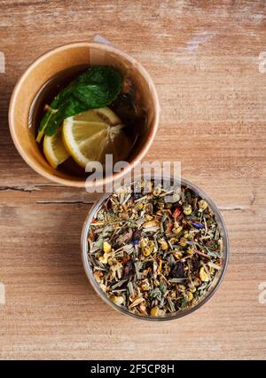 Bicchiere di foglie di tè e tè preparato con limone e menta in una tazza di carta monouso su sfondo rustico di legno. Vista dall'alto. Foto Stock