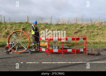 Un uomo guarda in un buco mentre installa cavi in fibra ottica su una strada rurale fuori Lisburn, Irlanda del Nord, Regno Unito Foto Stock