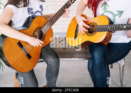 Bruna giovane in t-shirt con simbolo transgender seduto vicino Green Plant e suonare la chitarra spagnola contro il muro di mattoni Foto Stock