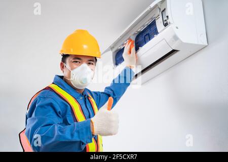 Happy asian maschio indossare una maschera di sicurezza per evitare che il tecnico della polvere pulisca il condizionatore d'aria all'interno, tecnico del condizionatore d'aria Foto Stock