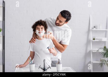 Sorridente padre arabo che applica la schiuma da barba sulla faccia della mensa figlio con rasoio in bagno Foto Stock