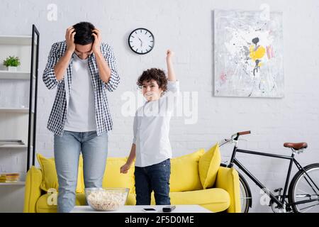 uomo arabo stanco che tocca la testa mentre si sta vicino a un figlio allegro mostrare il gesto di vittoria Foto Stock