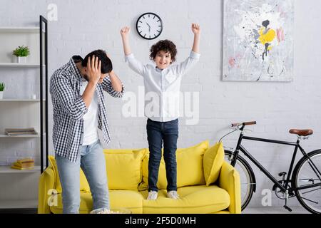 eccitato ragazzo arabo che salta sul divano vicino stanco padre copertura testa con le mani Foto Stock