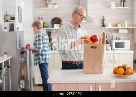Coppia senior in arrivo dal supermercato con borsa della spesa e disimballaggio in cucina. Anziani pensionati che si godono la vita, trascorrendo tempo ad aiutarsi l'un l'altro Foto Stock