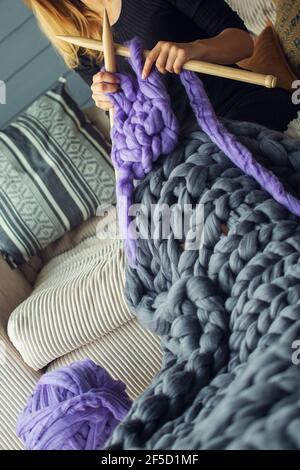 Una ragazza si siede su un divano del latte tesse un grigio e coperta di lana merino viola con aghi in legno Foto Stock