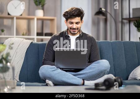 Giovane bell'uomo indiano o arabo che guarda una partita di calcio o un film interessante, seduto sul divano a casa con un PC portatile. Tempo libero a casa in serata. Foto Stock