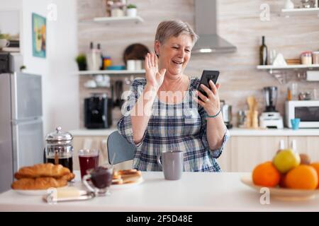 Donna anziana che saluta mentre videochiama con la famiglia utilizzando lo smartphone in cucina durante la colazione. Persona anziana che utilizza la tecnologia di chat online su Internet, webcam per tablet per chiamate in conferenza virtuali Foto Stock
