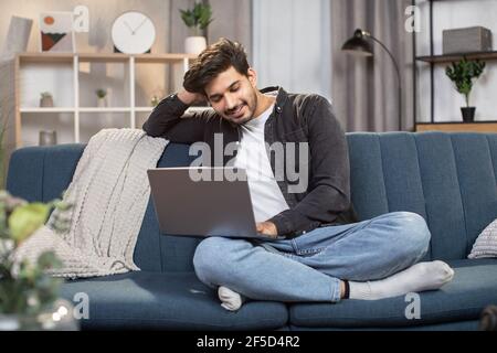 Positivo sorridente giovane indiano arabo in abiti casual, seduto sul morbido divano blu e chiacchierando con gli amici durante la videochiamata sul computer portatile mentre si passa il tempo a casa Foto Stock