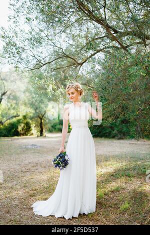 Una sposa tenera con un bouquet di fiori blu si erge da un olivo e tocca i rami con lei mano Foto Stock