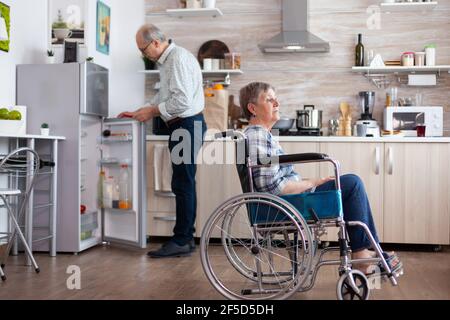 Donna anziana portatrice di handicap in sedia a rotelle seduta in cucina che guarda alla finestra mentre il marito disimballa la borsa della spesa. Vecchia signora disabile dopo lesione e riabilitazione, depresso non valido pieno di dolore Foto Stock