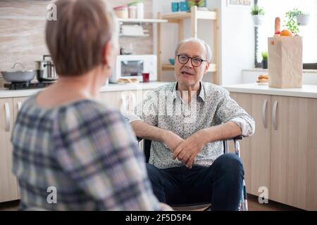 Uomo anziano disabile che parla con la moglie in cucina. Persona anziana che ha una conversazione con il marito in cucina. Vivere con persone disabili con disabilità motorie Foto Stock