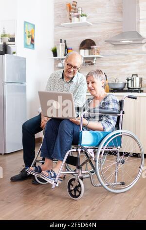 Donna anziana handicappata in sedia a rotelle e suo marito che cerca sul laptop, navigando sui social media seduti in cucina al mattino. Anziano paralizzato con una conferenza online. Foto Stock