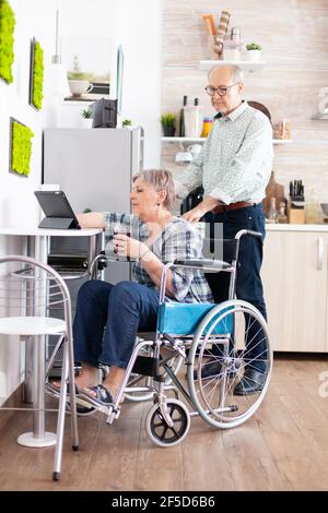 Donna anziana disabile in sedia a rotelle che utilizza un computer tablet in cucina con il marito vicino. Paralysied handicappato anziano persona utilizzando moderna comunicazione online internet web techonolgy. Foto Stock