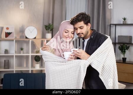 Famiglia, momenti accoglienti e concetto di amore. Carina coppia musulmana araba rilassante sotto coperta a casa nel soggiorno e bere caffè caldo Foto Stock