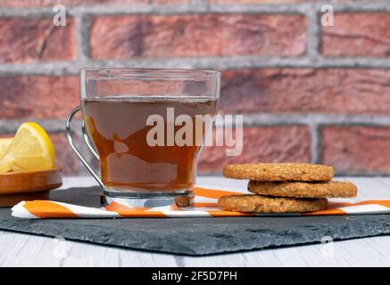 Biscotti d'avena su tovagliolo e piatto di pietra rettangolare, accompagnati da una tazza di tè e un piattino con fette di limone Foto Stock