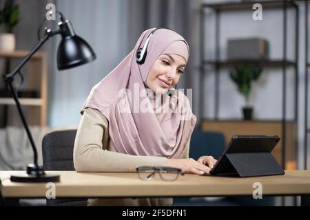 Concetto di lavoro a distanza da casa. Primo piano di una giovane donna arabica, attraente e sorridente, con foulard, che indossa le cuffie per lavorare da casa sul tablet pc Foto Stock