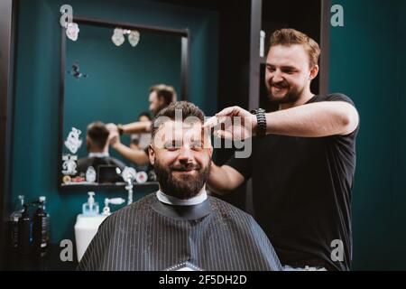 Felice giovane bearded cliente maschio di barbershop ottenere capelli mentre si siede in poltrona. Foto Stock