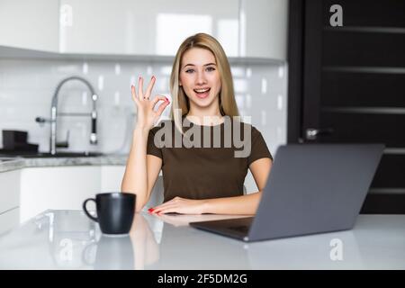 Giovane donna che usa il computer portatile in cucina facendo ok segno con dita Foto Stock