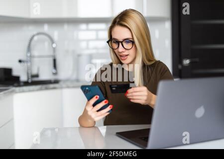 Ritratto indoor di donna seria attraente che tiene la carta di debito e il telefono delle cellule, inserendo i dati via il modulo di pagamento in linea mentre ordinando la consegna di cibo o s. Foto Stock