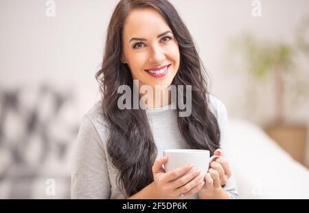 Giovane donna di brunetta che tiene una tazza di tè o caffè e che odora un aroma delizioso. Foto Stock