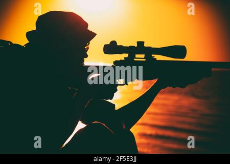 Il cecchino della squadra di Commando, lo sparatutto dell'esercito con forze speciali che punta, spara il fucile da cecchino mentre si siede sul mare o sulla riva dell'oceano durante il tramonto. Soldato di guardia costiera o di confine che osserva la costa con vista ottica Foto Stock