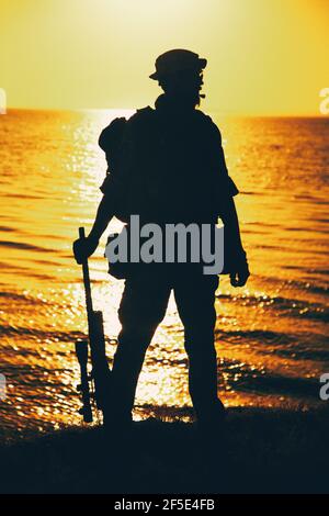 Silhouette di combattente comandante dell'esercito, cecchino della squadra Navy SEALS o soldato di guardia costiera in piedi sulla riva del mare con fucile sullo sfondo dell'orizzonte del tramonto sull'oceano. Forze speciali rifleman pattugliando costa Foto Stock