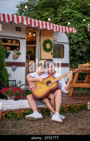 Il giovane padre con il suo piccolo figlio suonano la chitarra insieme di fronte alla loro casa rimorchio sullo sfondo a. una tavola di legno con una bella madre Foto Stock