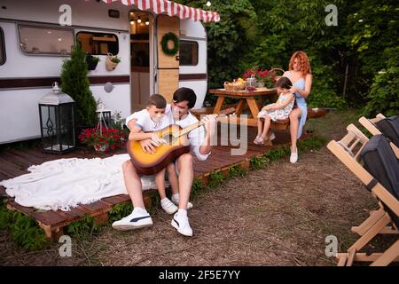 Il giovane padre con il suo piccolo figlio suonano la chitarra insieme di fronte alla loro casa rimorchio sullo sfondo a. una tavola di legno con una bella madre Foto Stock