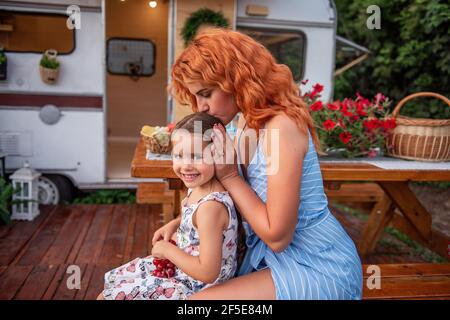 La giovane madre dai capelli rossi treccia le trecce di sua figlia piccola su panca di legno da camion rimorchio. Assistenza materna. La ragazza beve limonata. Picnic, campin Foto Stock