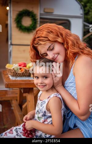 La giovane madre dai capelli rossi treccia le trecce di sua figlia piccola su panca di legno da camion rimorchio. Assistenza materna. La ragazza beve limonata. Picnic, campin Foto Stock