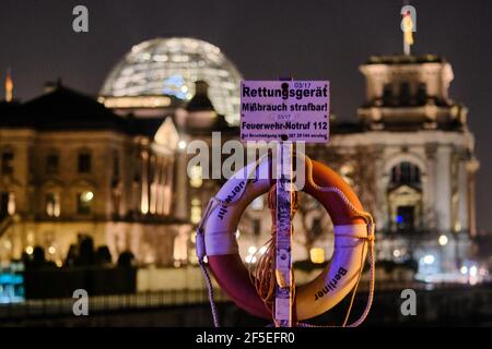 24 marzo 2021, Berlino: Un segno con l'iscrizione 'l'abuso di attrezzature di salvataggio punibile! Chiamata di emergenza antincendio 112' e un anello di vita stand sul Ponte Marschall, sullo sfondo l'edificio illuminato Reichstag e il Kronprinzenpalais (l), che ospita la società parlamentare, possono essere visti. Martedì sera (23 marzo 2021), i governi federale e statale avevano deciso, tra l'altro, di imporre un blocco pasquale più rigido dal 1 al 5 aprile per chiudere gravemente la vita pubblica, privata ed economica. A questo scopo, il giovedì e il sabato santo dovevano essere dichiarati giorni di riposo Foto Stock