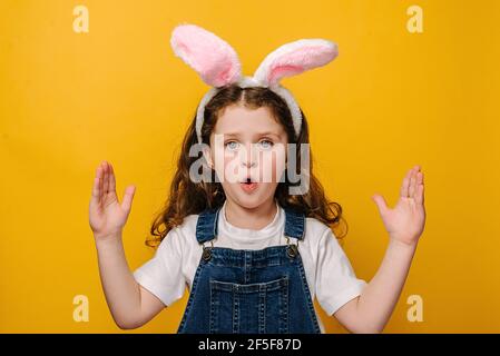 Scioccato eccitato bambino ragazza bella che indossa carino pink bunny orecchie fiducioso gesturing con mano facendo piccolo segno di dimensione, isolato su parete gialla Foto Stock