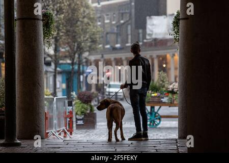 Un uomo e il suo cane si affacciano su una piazza deserta di Covent Garden durante una doccia a pioggia primaverile, nel centro di Londra, in Inghilterra, nel Regno Unito Foto Stock